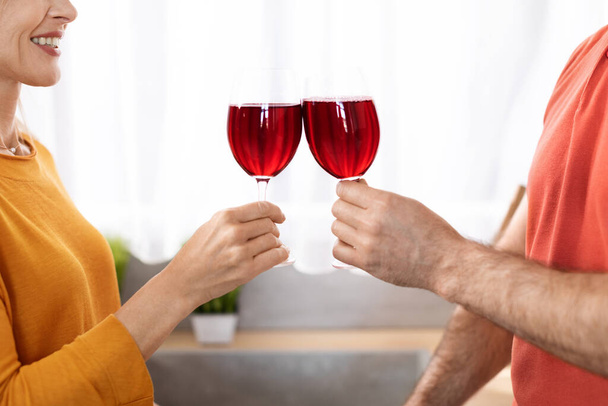 Εις υγείαν Αγνώριστο ζευγάρι γιορτάζει επέτειο στο σπίτι, τα χέρια του άνδρα και της γυναίκας κρατώντας ποτήρια με κόκκινο κρασί και επευφημίες, απολαμβάνοντας ρομαντική στιγμή μαζί, crop shot - Φωτογραφία, εικόνα
