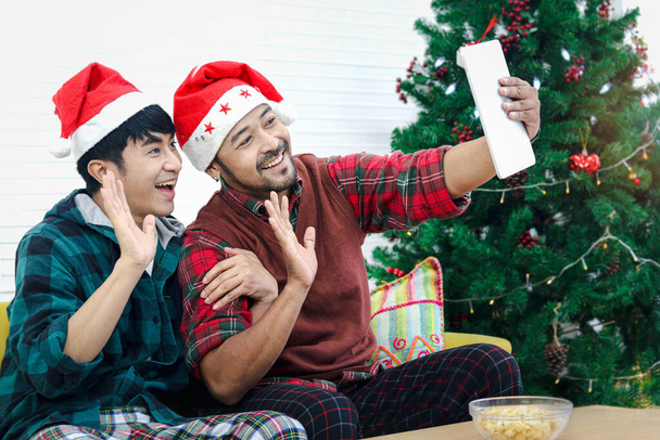 Cute happy young LGBT couple waving at digital tablet during video call for online greeting to family and friend, gay male lover sharing special moment on Christmas holiday night together. - Photo, Image