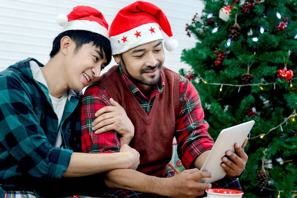 Cute happy young LGBT couple using digital tablet to video call for online greeting to family and friend, gay male lover sharing special moment on Christmas holiday night together. - Photo, Image