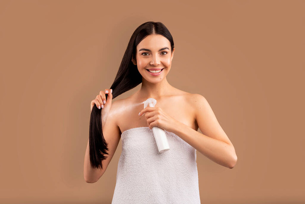 Lächelnde halbnackte langhaarige brünette junge Frau in weißes Badetuch gehüllt mit Haarspray nach der Dusche vor dem Styling, isoliert auf beigem Studiohintergrund, Kopierraum. Haarstyling-Konzept - Foto, Bild