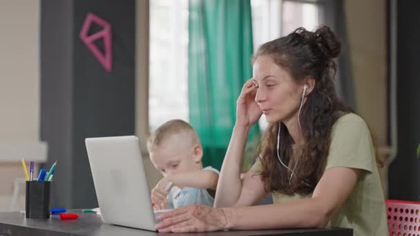 pracująca mama i jej małe dziecko w mieszkaniu, kobieta pracuje online przez laptopa, ładny chłopiec gra w pobliżu, samotna matka na urlopie macierzyńskim, współczesne kobiety z dziećmi, szczęśliwa rodzina - Materiał filmowy, wideo