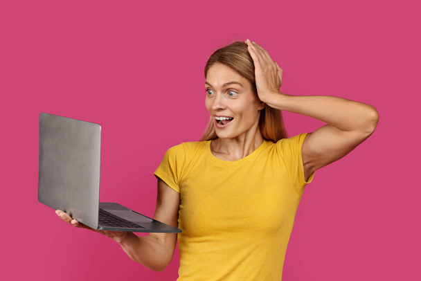 Smiling surprised adult caucasian blonde lady in yellow t-shirt with open mouth holds hand at head looks at laptop, isolated on pink background. Offer and ad, online win, success, huge sale, emotions - Photo, Image