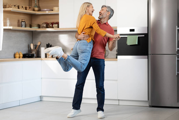 Loving middle aged grey-haired handsome man and pretty blonde woman in casual outfit enjoying time together at home, dancing in kitchen, husband lifting up laughing wife, full length, copy space - Photo, Image