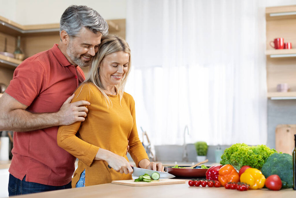 Loving handsome husband middle aged man in casual hugging his smiling attractive spouse at cozy kitchen, mature woman cooking healthy delicious dinner for her family at home, copy space - Foto, Bild