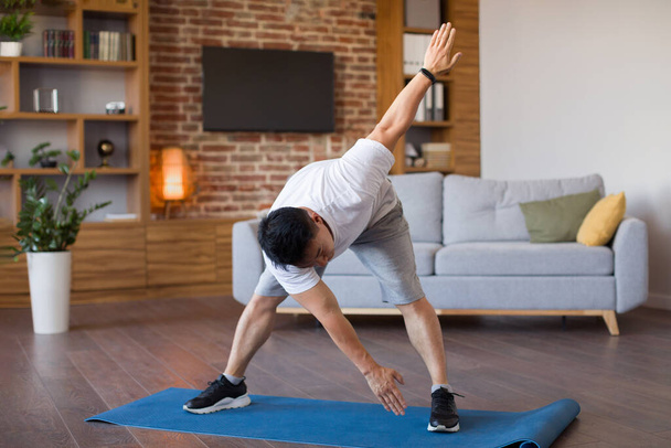 Melegíts be és nyújtózkodj edzés közben. Aktív érett ázsiai férfi dőlés és nyújtás a nappaliban belső, álló fitness mat, szabad hely - Fotó, kép