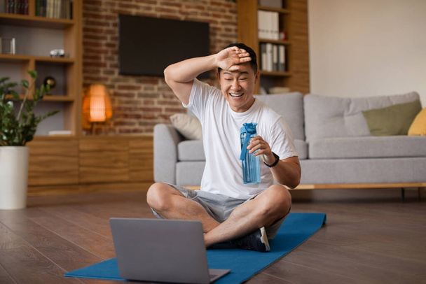 Onnellinen väsynyt aasialainen mies tunne uupunut jälkeen online harjoitus, käyttäessään kannettavaa tietokonetta, istuu maton kotona. Urheilu aloittelija sturuggling treenata sisätiloissa - Valokuva, kuva