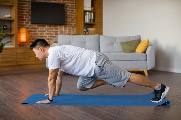 Active mature asian man doing cross body mountain climbers exercises on fitness mat, training in living room interior, copy space, side view - Photo, Image