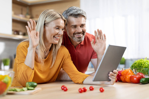Cheerful attractive mature man and woman in homewear cooking and having video call with friends or kids, food vloggers streaming, using digital tablet at kitchen, waving at pad screen, copy space - Photo, Image