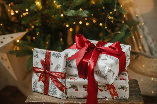 Stílusos karácsonyi ajándékok karácsonyfán fényekkel. Boldog Karácsonyt és Kellemes Ünnepeket! Csomagolt karácsonyi ajándékok piros szalaggal díszített fa alatt a hangulatos szobában. Légköri transzparens - Fotó, kép