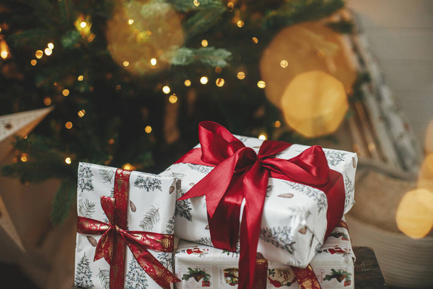 Frohe Weihnachten und frohe Feiertage! Stilvolle Weihnachtsgeschenke am Weihnachtsbaum mit goldenen Lichtern bokeh. Eingewickelte Weihnachtsgeschenke mit roter Schleife unter geschmücktem Baum im Zimmer. Stimmungsvolles Banner - Foto, Bild