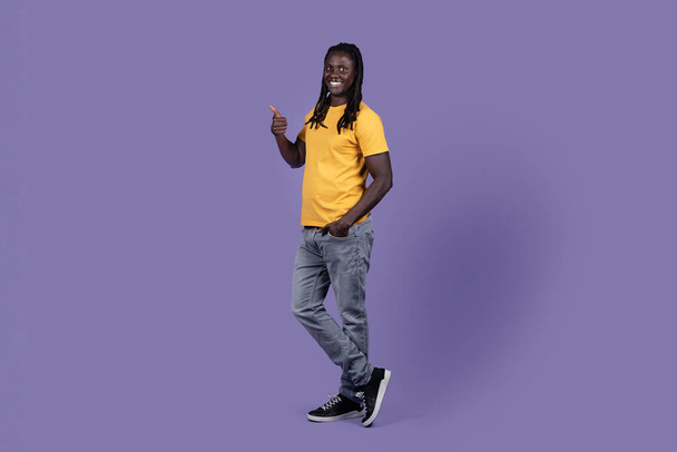 Cheerful attractive african american guy with long dreadlocks in casual outfit showing nice offer over purple studio background, pointing with thumb up at copy space and smiling, full length shot - Photo, Image