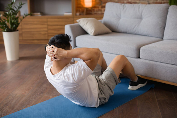 Активный взрослый человек осуществляет мышцы пресса на коврике йоги возле дивана в доме, в полную длину. Подходят мужчины, ведущие здоровый образ жизни, тренирующиеся во время тренировок, занимающиеся домашним фитнесом - Фото, изображение