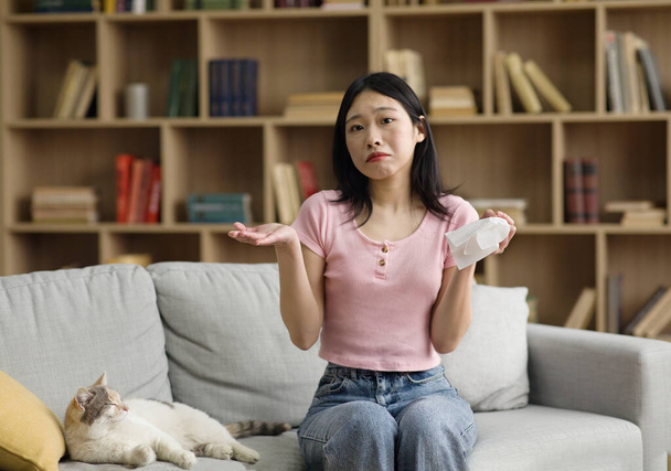 Rozrušená korejská dáma držící papírovou tkáň, která má alergii na srst a trpí rýma způsobenou kocourem, sedí doma na pohovce, kopíruje prostor - Fotografie, Obrázek