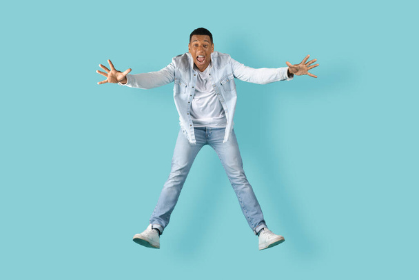 Surprised excited amazed happy young african american man with open mouth screaming, has fun, jumped, frozen in air, isolated on blue background, studio. Guy enjoys fly, free time, huge ad and offer - Фото, зображення