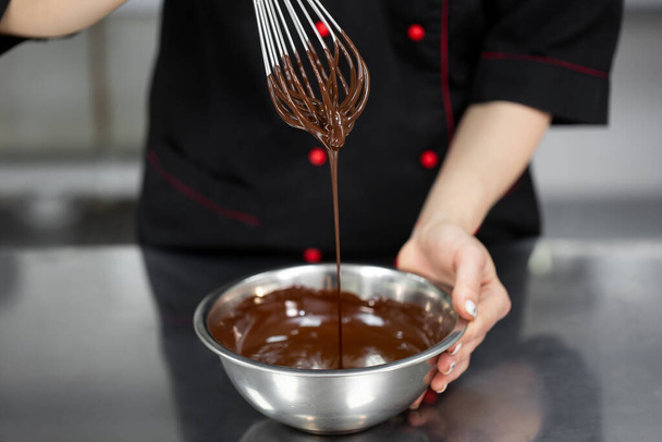 Pastry chef whips melted chocolate in a bowl with a metal wire whisk close up - Photo, Image