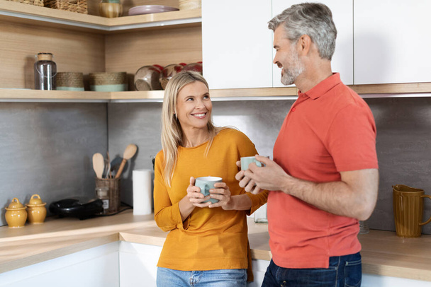 Loving beautiful middle aged couple drinking coffee at home, happy mature husband and wife in comfy homewear standing at cozy kitchen, holding tea mugs, looking at each other and smiling, copy space - Photo, Image