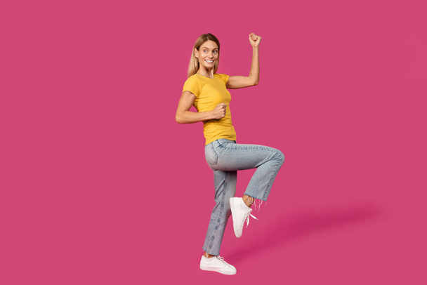 Cheerful middle aged caucasian blonde lady in yellow t-shirt making victory and success gesture, isolated on pink background, full length, studio. Win sing, advertising and offer and people emotions - Fotó, kép