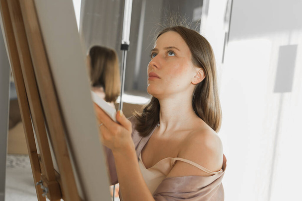 Νεαρή γυναίκα καλλιτέχνης ζωγραφική σε καμβά στο καβαλέτο στο σπίτι στο υπνοδωμάτιο - τέχνη και δημιουργικότητα - Φωτογραφία, εικόνα