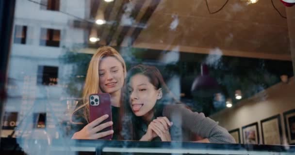 Młode kobiety robiące selfie w restauracji. Niski kąt uchwyt młody brunetka pokazując język podczas robienia selfie ze szczęśliwą dziewczyną za szkłem w restauracji - Materiał filmowy, wideo