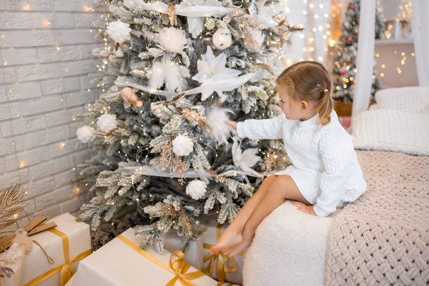 Muy bonita niña encantadora rubia en vestido blanco sentada en una cama infantil en el fondo de los árboles de Navidad en el interior brillante de la casa. Foto de alta calidad - Foto, imagen