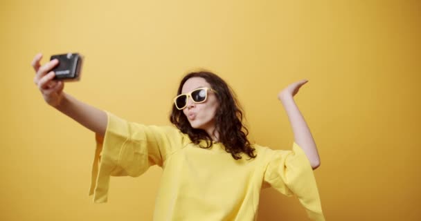 Stylish young woman taking selfie. Static shot of happy young female pouting lips and gesturing V sign then showing tongue and sending air kiss while taking selfie against yellow background - Footage, Video