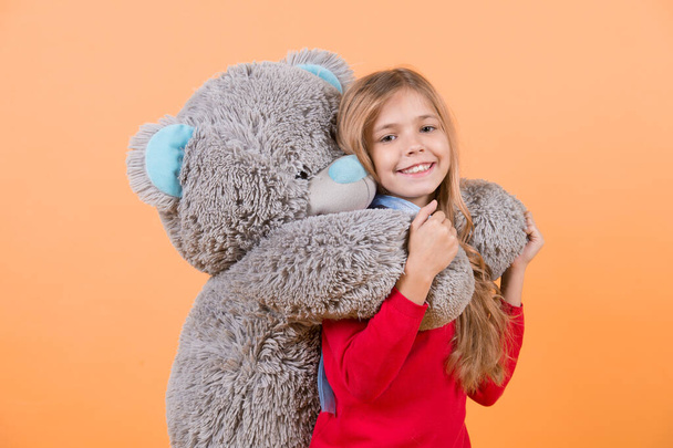 Happy childhood concept. Kid with animal doll, present and gift. Child smile with grey soft toy. Girl hug big teddy bear on orange background. Holiday, birthday, anniversary celebration. - Photo, Image