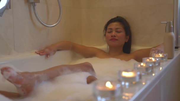 Femme détendue avec les yeux fermés profitant d'une baignoire pétillante entourée de bougies. Bougies lumineuses alignées pour une atmosphère relaxante et une expérience agréable tout en ayant un traitement spa dans la salle de bain à la maison. - Séquence, vidéo