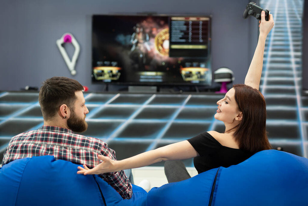 Young couple playing video games, sitting on chairs in a gaming club with controllers in their hands, a rear view from the TV screen. - Foto, imagen