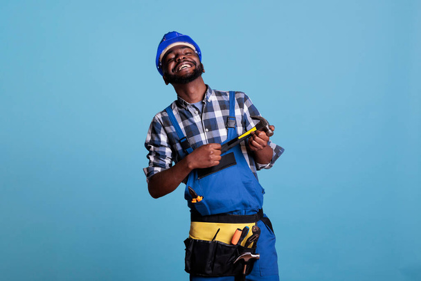 Studio photo of glad african american builder in helmet and utility belt holding hammer with two hands. Happy construction worker smiling proudly on blue background in studio shot. - Photo, Image