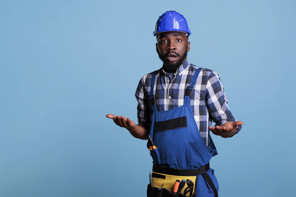 Ritratto di un confuso costruttore afroamericano che posa in uniforme da lavoro su sfondo blu, rendendo incerta l'espressione facciale. Uomo che scuote le spalle, colpo in studio Non so concetto. - Foto, immagini