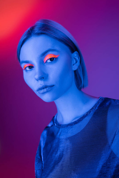 young woman with glowing makeup in blue neon light on purple and coral background - Foto, Imagen