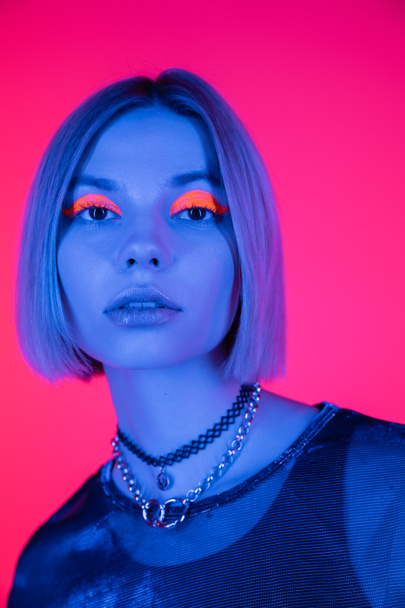 portrait of young woman with neon makeup looking at camera in blue light isolated on deep pink - Фото, зображення