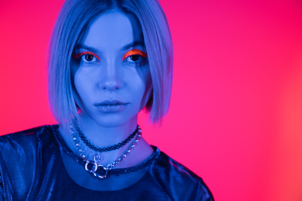 portrait of trendy woman with necklaces and fluorescent makeup in blue neon light on deep pink background - Foto, Imagen