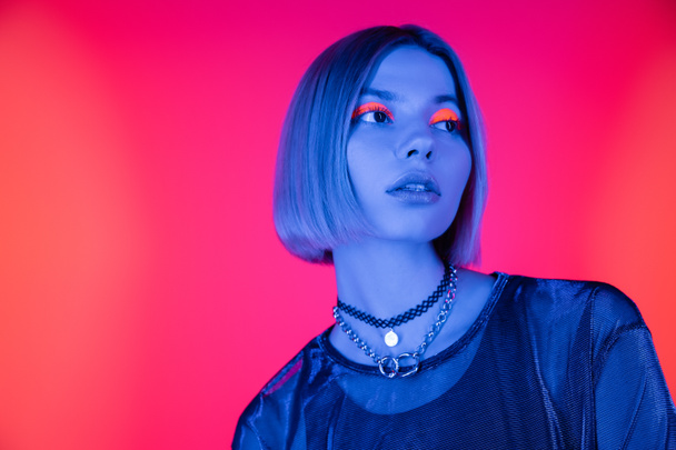 fashionable woman with neon makeup looking away in blue light on coral pink background - 写真・画像