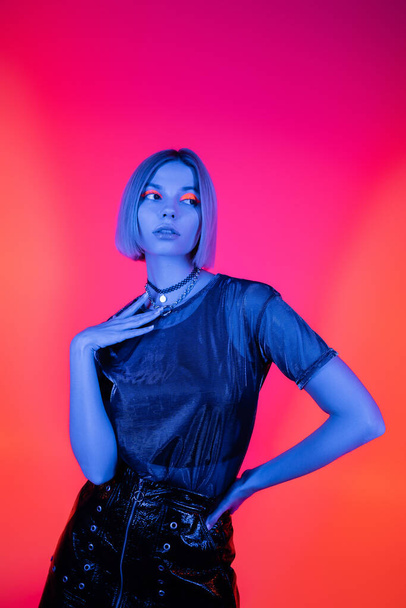 young and trendy woman posing with hand on hip in blue neon light on coral and pink background  - Fotografie, Obrázek