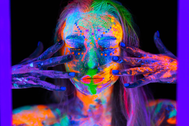 Beautiful young woman in neon light. Portrait of a model with fluorescent makeup posing in UV light with colorful makeup - 写真・画像