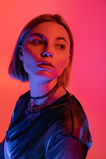 portrait of woman in necklaces looking away in neon light on pink and coral background - Foto, imagen