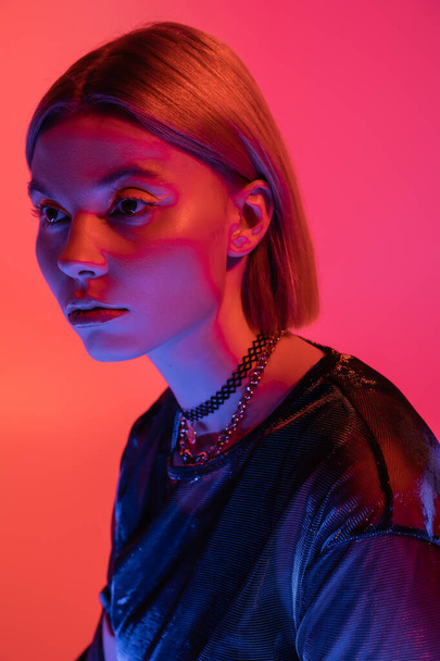 portrait of trendy woman in necklaces posing in neon light on coral and pink background - Foto, afbeelding