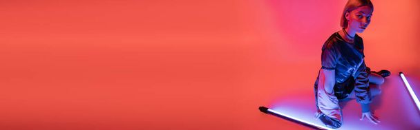 trendy woman sitting near purple fluorescent lamps on coral background, banner - Photo, Image