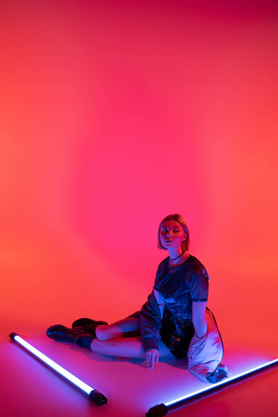 полная длина стильная женщина сидит рядом с фиолетовыми неоновыми лампами на коралловом красном фоне - Фото, изображение