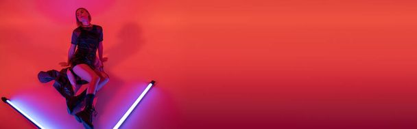 vista ad alto angolo di giovane donna in stivali e minigonna posa vicino brillanti lampade al neon su sfondo viola e rosso, banner - Foto, immagini