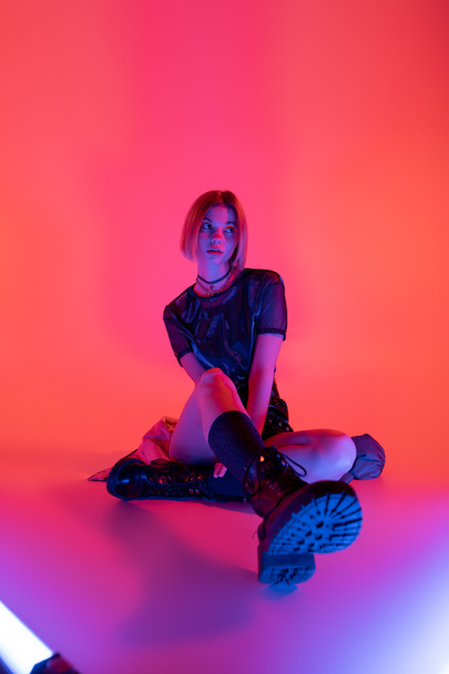young woman in stylish leather boots looking away while sitting in neon light on purple and coral background - Фото, зображення