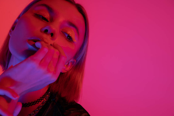 ritratto di giovane donna con trucco al neon che tiene la mano vicino al viso su sfondo rosa scuro - Foto, immagini
