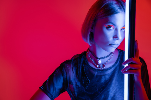 stylish woman in bright makeup and necklaces looking at camera in blue light of neon lamp on carmine red background - Photo, Image