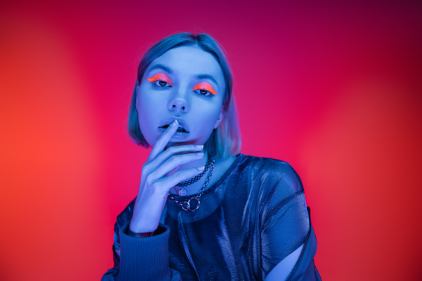 sensual young woman with neon makeup touching lips in blue light on coral and pink background - Фото, изображение