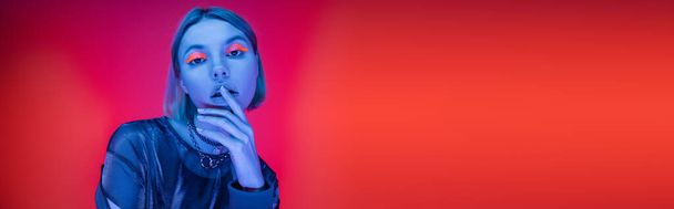 mulher elegante com maquiagem de néon tocando lábios em luz azul no fundo vermelho coral, banner - Foto, Imagem