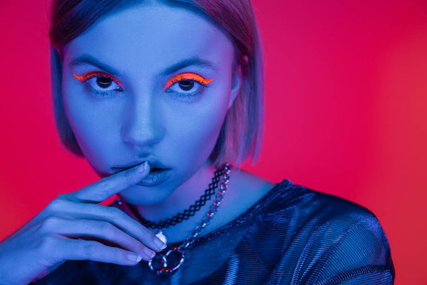 sensual woman with glowing makeup touching lips in blue neon light on coral pink background - Фото, зображення