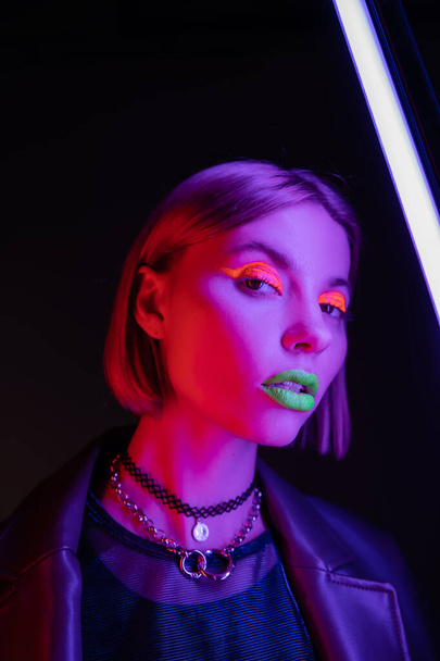 mulher na moda com maquiagem neon brilhante olhando para a câmera perto de lâmpada fluorescente roxa no fundo preto - Foto, Imagem