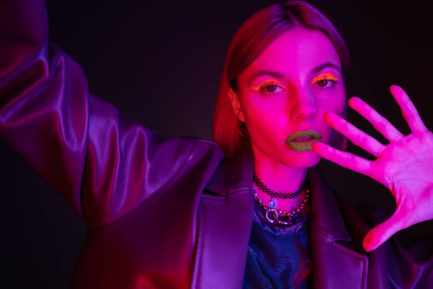 žena v kožené bundě a pulzující neon make-up při pohledu na kameru na tmavém fialovém pozadí - Fotografie, Obrázek