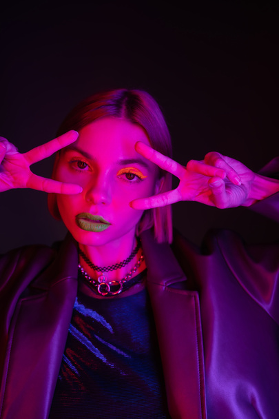mulher na moda em maquiagem de néon e colares mostrando sinais de vitória e olhando para a câmera em luz roxa no fundo preto - Foto, Imagem